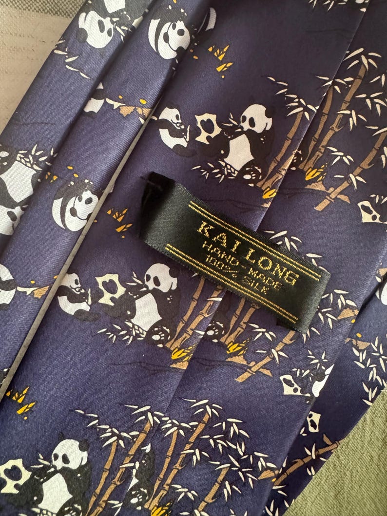 May include: A dark blue necktie featuring a repeating pattern of pandas eating bamboo stalks. A black fabric label is stitched onto the front, displaying the gold text KAI LONG, HAND-MADE, and 100% SILK in a clear, legible font.