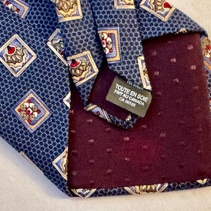 May include: A blue necktie featuring a repeating pattern of squares containing floral motifs in red, white, and tan. The back of the tie has a dark maroon lining with a subtle dot texture and a label reading TOUTE EN SOIE FAIT AU CANADA CA 00125.