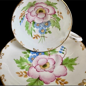 May include: A white teacup and saucer set with a floral design. The cup and saucer feature pink and blue flowers with green leaves and gold accents.