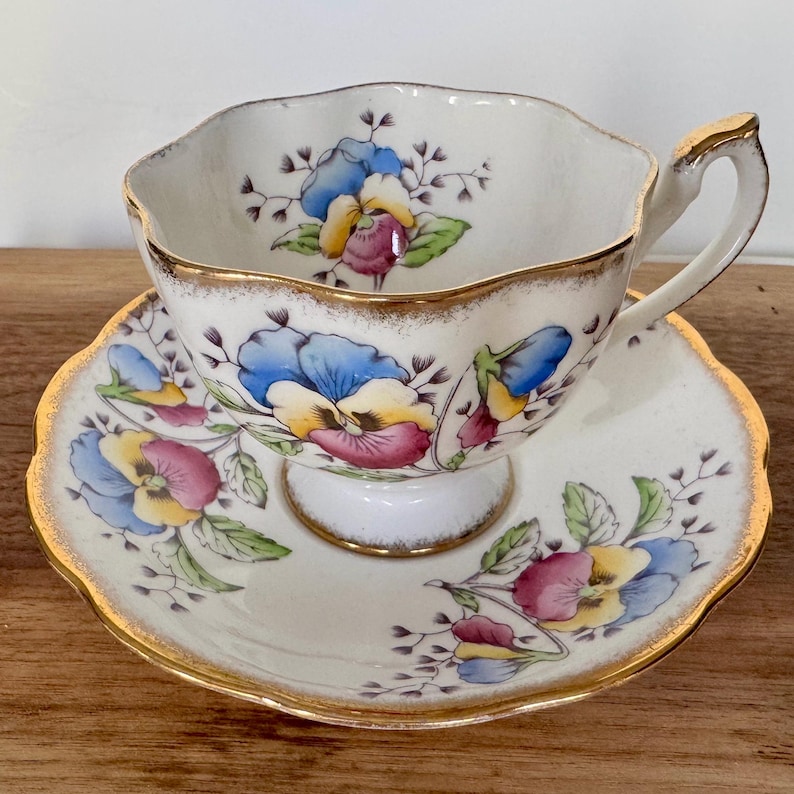 May include: A vintage teacup and saucer set. The white porcelain cup and saucer feature a floral design with blue, yellow, and purple pansies. Gold trim accents the scalloped edges of the cup and saucer, and the handle of the cup.