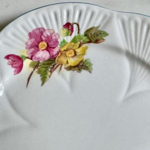 May include: White ceramic plate with a scalloped edge and a floral design. The flowers are pink and yellow with green leaves. The plate has a ridged texture and a light blue trim.