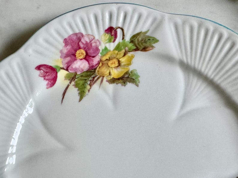 May include: White ceramic plate with a scalloped edge and a floral design. The flowers are pink and yellow with green leaves. The plate has a ridged texture and a light blue trim.