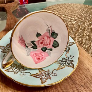 May include: A vintage teacup and saucer set with a delicate floral design. The cup features a white background with pink roses and green leaves. The saucer is a light blue color with gold accents and a leafy pattern.