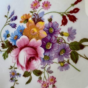 May include: Close-up of a white ceramic plate with a vibrant floral design. The arrangement features a large pink rose, purple, blue, and orange flowers, and green leaves. The plate has a gold trim.