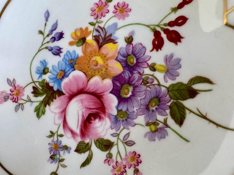 May include: Close-up of a white ceramic plate with a vibrant floral design. The arrangement features a large pink rose, purple, blue, and orange flowers, and green leaves. The plate has a gold trim.