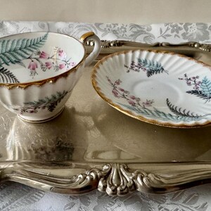 Puede incluir: Un juego de taza y platillo de t&eacute; vintage con un dise&ntilde;o floral de helecho en verde, rosa y dorado. La taza y el platillo est&aacute;n sobre una bandeja plateada. La taza tiene un asa y un borde dorados. El platillo tiene un borde dorado.