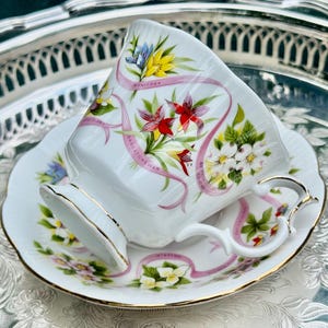 May include: White teacup and saucer set with gold trim, featuring floral and ribbon designs. The cup and saucer are decorated with colorful flowers and the text "Manitoba". The set is placed on a silver tray.