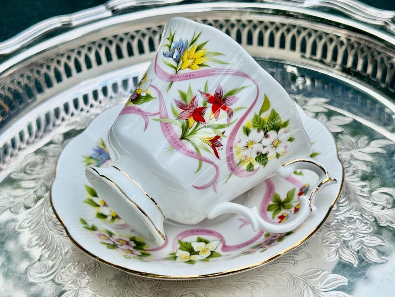 May include: White teacup and saucer set with gold trim, featuring floral and ribbon designs. The cup and saucer are decorated with colorful flowers and the text "Manitoba". The set is placed on a silver tray.