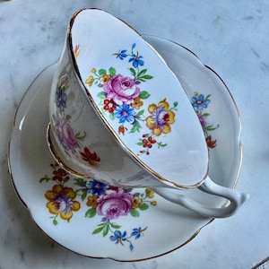 Puede incluir: Juego de taza y platillo blancos con dise&ntilde;o floral. Presenta rosas rosas, flores amarillas y azules, y ribete dorado. La taza est&aacute; inclinada, mostrando el interior. El platillo tiene un borde festoneado.