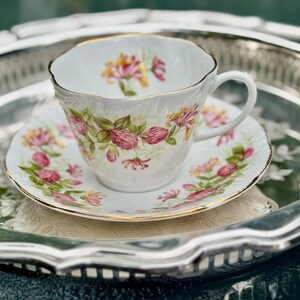 May include: A white teacup and saucer set with a floral design in pink, yellow, and green, with gold trim. The teacup and saucer are on a silver tray, creating an elegant tea set.