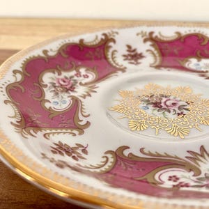 May include: Close-up of a white and pink porcelain saucer with gold trim. The saucer features a floral design with gold filigree accents and a scalloped edge. A decorative piece for serving tea or coffee.