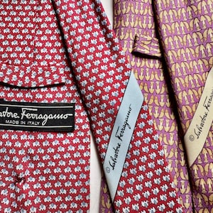 Puede incluir: Dos corbatas Salvatore Ferragamo. Una es roja con un patr&oacute;n repetitivo de elefantes blancos y una etiqueta negra. La otra es morada con un patr&oacute;n abstracto dorado repetitivo y una etiqueta color crema. Ambas muestran la marca.