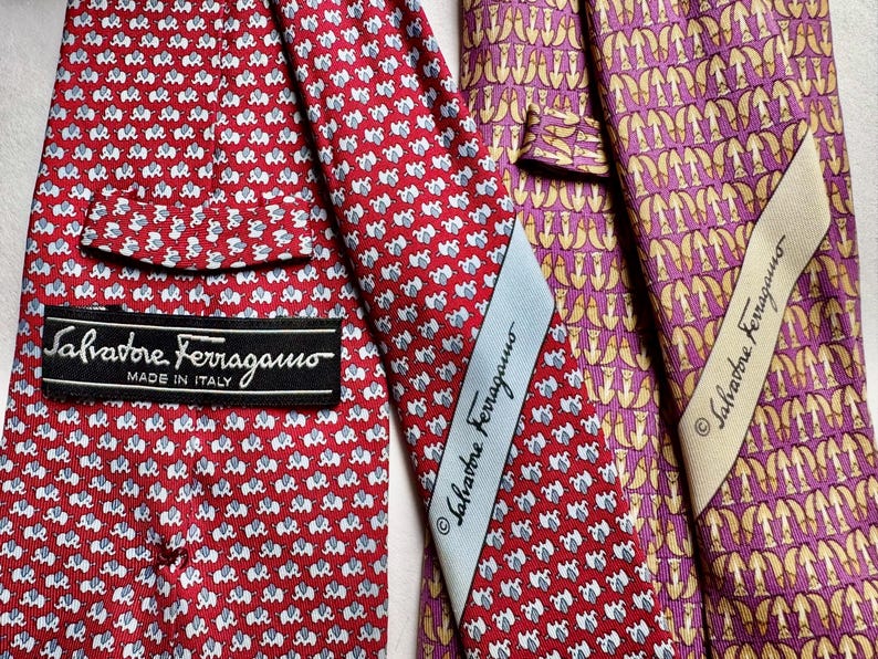 Puede incluir: Dos corbatas Salvatore Ferragamo. Una es roja con un patr&oacute;n repetitivo de elefantes blancos y una etiqueta negra. La otra es morada con un patr&oacute;n abstracto dorado repetitivo y una etiqueta color crema. Ambas muestran la marca.