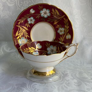 May include: A porcelain teacup and saucer set featuring a deep burgundy background with light blue floral motifs and gold leaf accents. The teacup has a scalloped rim, a gold-trimmed handle, and a gold base, while the saucer matches the design.