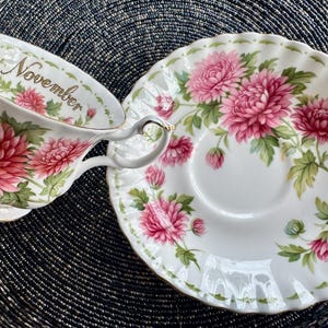 May include: White teacup and saucer set with a floral design. The teacup has a handle and the word "November" printed on the rim. The floral design features pink flowers and green leaves. The saucer has a matching floral pattern.
