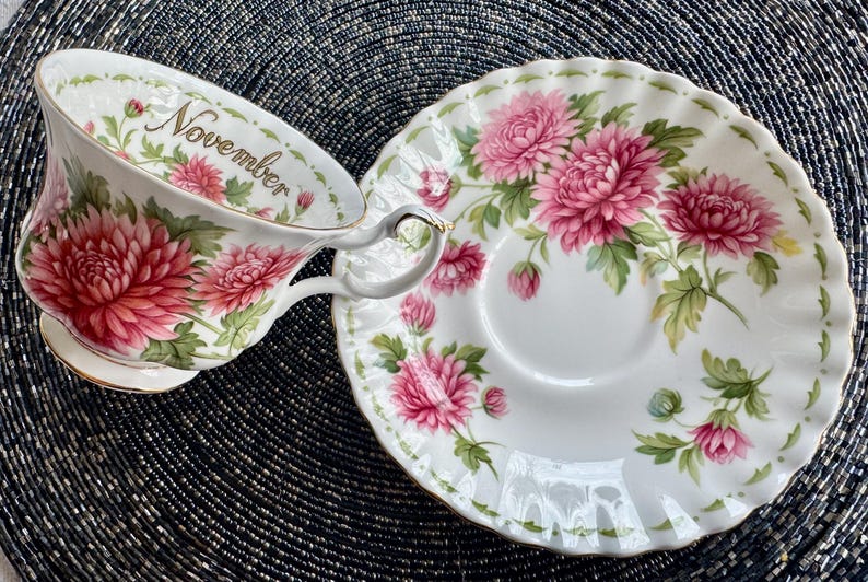 May include: White teacup and saucer set with a floral design. The teacup has a handle and the word "November" printed on the rim. The floral design features pink flowers and green leaves. The saucer has a matching floral pattern.