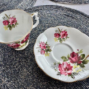 Puede incluir: Un juego de taza y platillo de t&eacute; blanco con un patr&oacute;n floral con rosas rosas y rojas, hojas verdes y detalles dorados. El dise&ntilde;o floral est&aacute; sobre un fondo blanco.