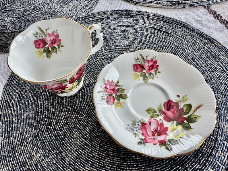 Puede incluir: Un juego de taza y platillo de t&eacute; blanco con un patr&oacute;n floral con rosas rosas y rojas, hojas verdes y detalles dorados. El dise&ntilde;o floral est&aacute; sobre un fondo blanco.