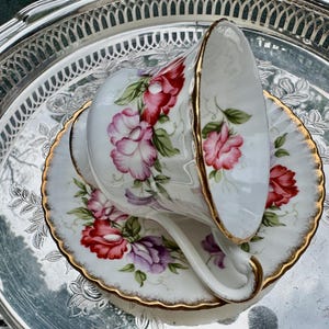 May include: A white teacup and saucer set with a floral rose design in red, pink, and purple, accented with gold trim. The teacup is resting on a silver tray, creating an elegant presentation.