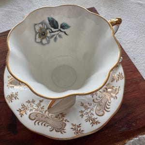 May include: A white teacup and saucer with a gold rim and floral design. The teacup has a single white flower with a green stem and leaves. The saucer has a delicate gold pattern.