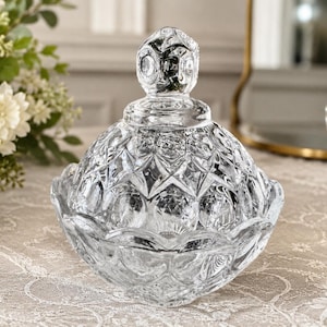 May include: A clear glass candy dish with a matching lid, featuring a faceted diamond and circular pattern across the surface. The lid is topped with a decorative finial, and the bowl has a scalloped edge, reflecting light through the glass.