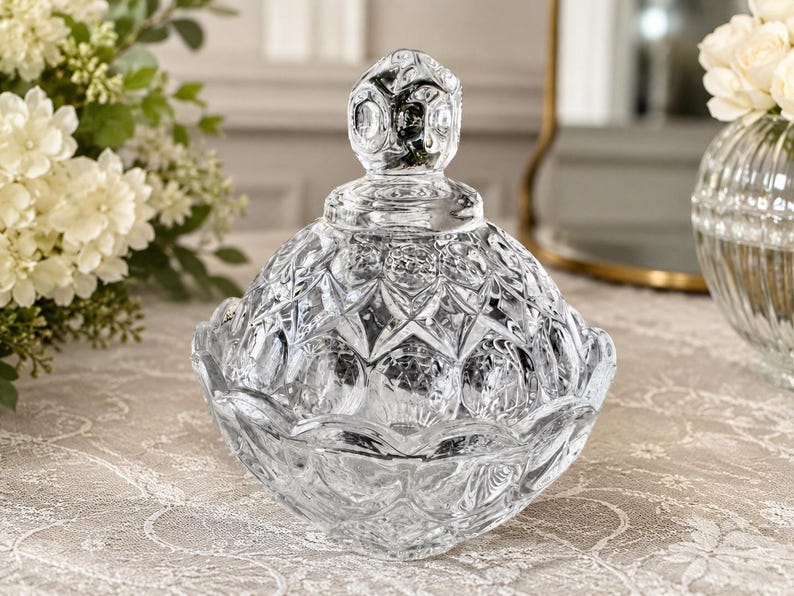 May include: A clear glass candy dish with a matching lid, featuring a faceted diamond and circular pattern across the surface. The lid is topped with a decorative finial, and the bowl has a scalloped edge, reflecting light through the glass.