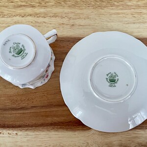 May include: White Bone China teacup and saucer set. The teacup has a handle and floral design. The saucer has a scalloped edge. Both pieces have a green emblem and text that reads "Bone China, Hammersley, Made in England."
