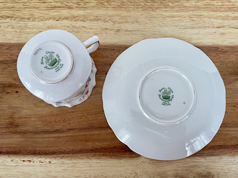May include: White Bone China teacup and saucer set. The teacup has a handle and floral design. The saucer has a scalloped edge. Both pieces have a green emblem and text that reads "Bone China, Hammersley, Made in England."