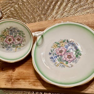 May include: A set of two white porcelain teacups with a green and gold rim. The cups feature a floral design with pink, yellow, and blue flowers.