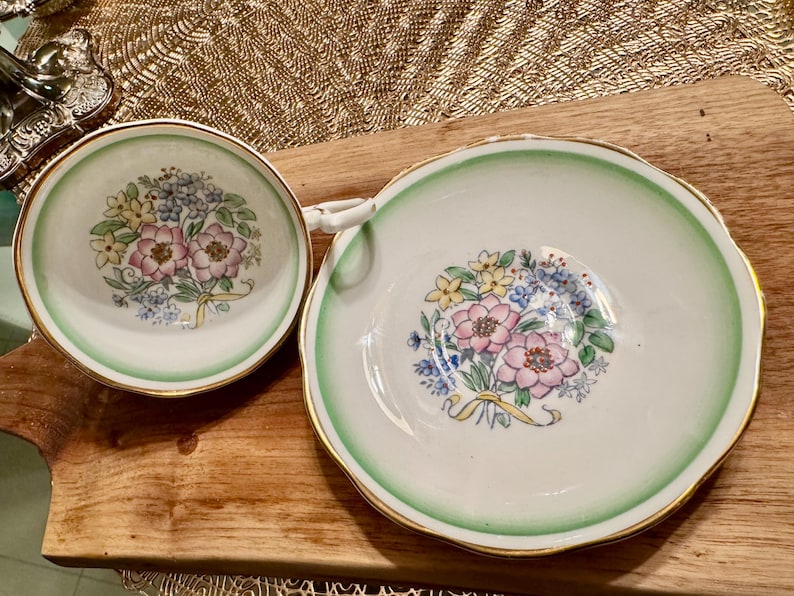 May include: A set of two white porcelain teacups with a green and gold rim. The cups feature a floral design with pink, yellow, and blue flowers.
