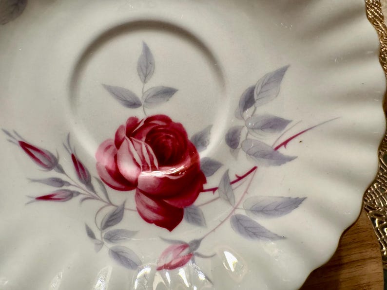 May include: A white porcelain saucer with a scalloped edge features a hand-painted design of a single red rose with gray leaves. The saucer is perfect for tea or coffee.