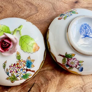 May include: A porcelain trinket box with a floral design and a butterfly. The lid features a pink rose with green leaves and a butterfly with yellow, blue, and white wings. The bottom of the box has a gold rim and a blue and white Herend Hungary logo.