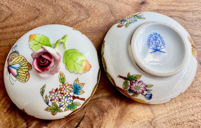 May include: A porcelain trinket box with a floral design and a butterfly. The lid features a pink rose with green leaves and a butterfly with yellow, blue, and white wings. The bottom of the box has a gold rim and a blue and white Herend Hungary logo.