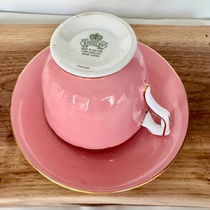 May include: A pink teacup and saucer set with gold trim. The teacup handle is white with gold accents. The saucer and teacup are a solid pink color. The bottom of the teacup has the text "AYNSLEY MADE IN ENGLAND FINE ENGLISH BONE CHINA".
