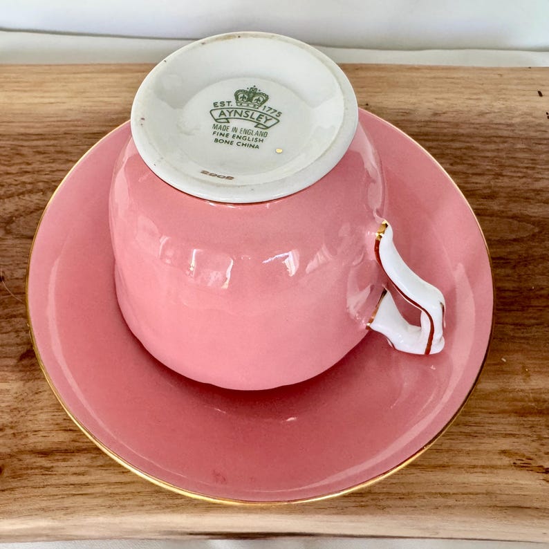 May include: A pink teacup and saucer set with gold trim. The teacup handle is white with gold accents. The saucer and teacup are a solid pink color. The bottom of the teacup has the text "AYNSLEY MADE IN ENGLAND FINE ENGLISH BONE CHINA".