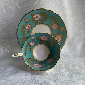 May include: A turquoise teacup and matching saucer set featuring a white center, scalloped edges with gold trim, and a floral pattern of pink blossoms with gold leaves. The teacup handle is white with a small gold leaf detail on the top.