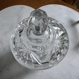 May include: A clear glass candy dish with a matching lid featuring a faceted knob handle. The lid is decorated with a pressed starburst pattern radiating from the center, creating a reflective surface with sharp geometric lines and clear glass.