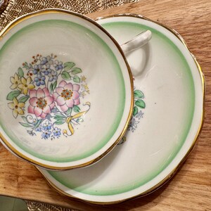 May include: A vintage teacup and saucer set with a floral design. The cup and saucer are white with a green rim and gold trim. The floral design features pink, yellow, and blue flowers.