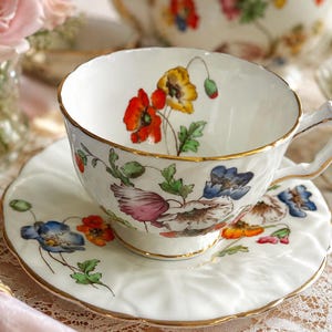 K&ouml;nnte beinhalten: Wei&szlig;es Tee-Set mit floralem Design, darunter rote, gelbe und blaue Blumen. Die Tasse und Untertasse haben einen Goldrand. Das Set steht auf einer Spitzendecke, im Hintergrund eine Vase mit rosa Rosen.