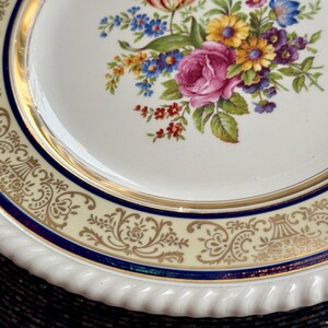 May include: A white porcelain plate with a floral design. The plate features a gold rim and a blue and gold patterned border. The center of the plate is decorated with a bouquet of pink roses, yellow daisies, and blue flowers.