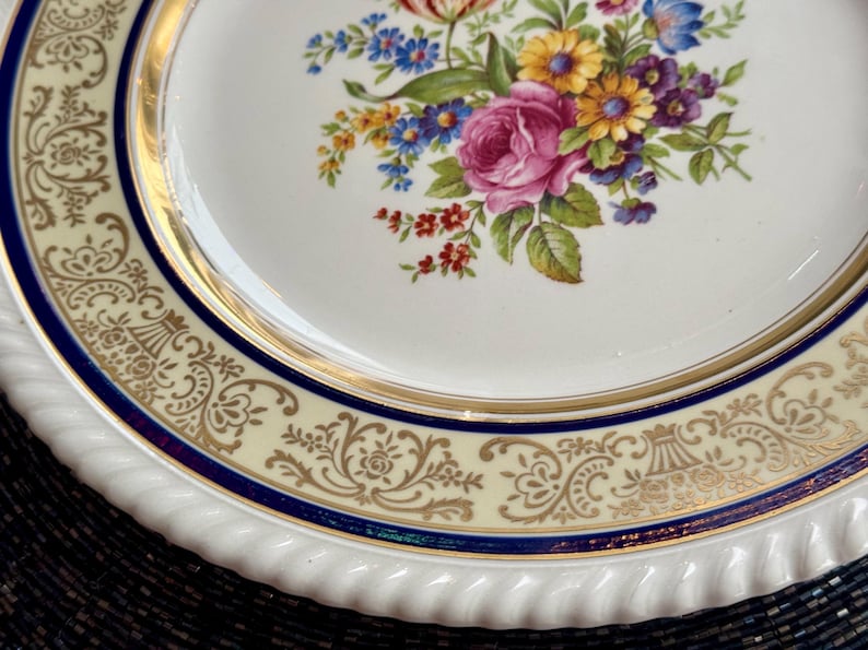 May include: A white porcelain plate with a floral design. The plate features a gold rim and a blue and gold patterned border. The center of the plate is decorated with a bouquet of pink roses, yellow daisies, and blue flowers.