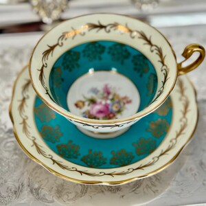 May include: A vintage teacup and saucer set. The teacup interior and saucer are teal, with gold floral accents and a floral center design. The rim and handle are gold, and the outer rim is cream-colored.