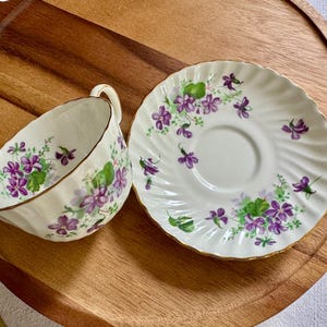 K&ouml;nnte beinhalten: Ein wei&szlig;es Tee-Set mit einer Tasse und Untertasse mit einem violetten Blumendesign. Die Tasse und Untertasse haben einen goldenen Rand. Das Set befindet sich auf einem Holztablett.