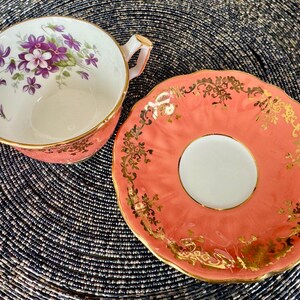 May include: A coral pink teacup and saucer set with a floral design and gold trim. The teacup has a white interior and a floral design with purple flowers. The saucer has a scalloped edge and a gold floral design.
