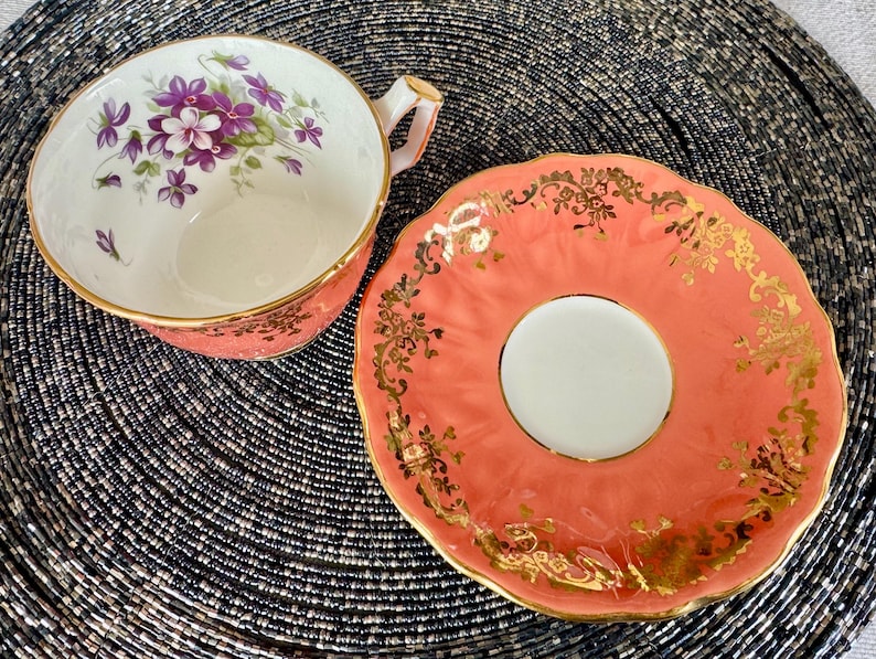 May include: A coral pink teacup and saucer set with a floral design and gold trim. The teacup has a white interior and a floral design with purple flowers. The saucer has a scalloped edge and a gold floral design.
