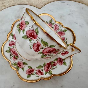 Puede incluir: Juego de taza y platillo blancos con un dise&ntilde;o floral de flores rosas y bordes dorados. La taza est&aacute; apoyada de lado sobre el platillo a juego. Un dise&ntilde;o cl&aacute;sico.