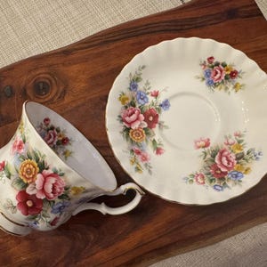 May include: A white teacup and saucer with a floral pattern. The cup and saucer have a delicate, scalloped edge. The floral pattern features pink, red, yellow, and blue flowers.
