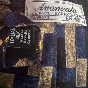 May include: A necktie featuring a geometric pattern in navy blue, black, and gold. A white label reads Avanzata FOR/POUR HARRY ROSEN CA 00572 RN 76912. A black label reads ITALIAN SILK MADE IN CANADA CA 00952. The fabric has a textured, ribbed appearance.