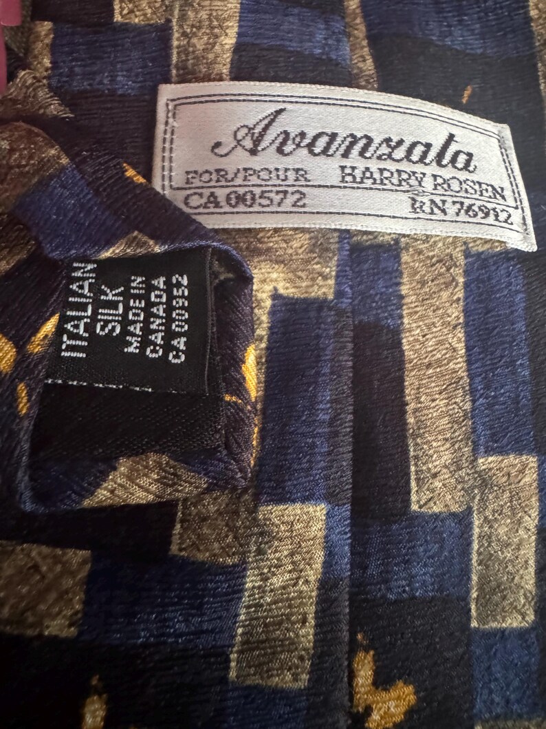 May include: A necktie featuring a geometric pattern in navy blue, black, and gold. A white label reads Avanzata FOR/POUR HARRY ROSEN CA 00572 RN 76912. A black label reads ITALIAN SILK MADE IN CANADA CA 00952. The fabric has a textured, ribbed appearance.