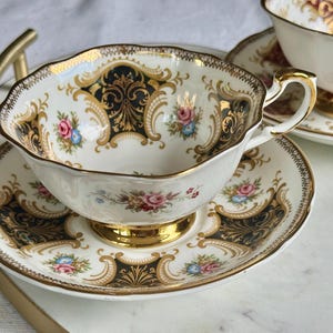 May include: A white teacup and saucer set with a gold rim and handle. The cup and saucer feature a black and gold ornate design with pink and blue floral accents. The set is placed on a white surface.